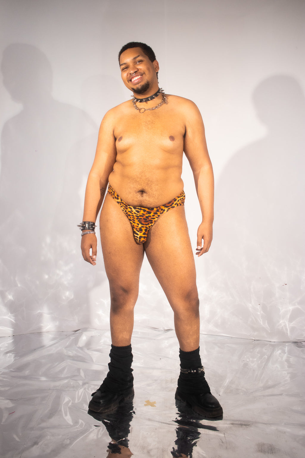 A man modeling a low-rise leopard print thong-style brief with black socks and black boots, standing against a white studio backdrop with a reflective floor.