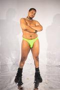 A man posing in bright neon green thong underwear with a pink and white pattern, styled with black boots and accessories against a white studio backdrop.