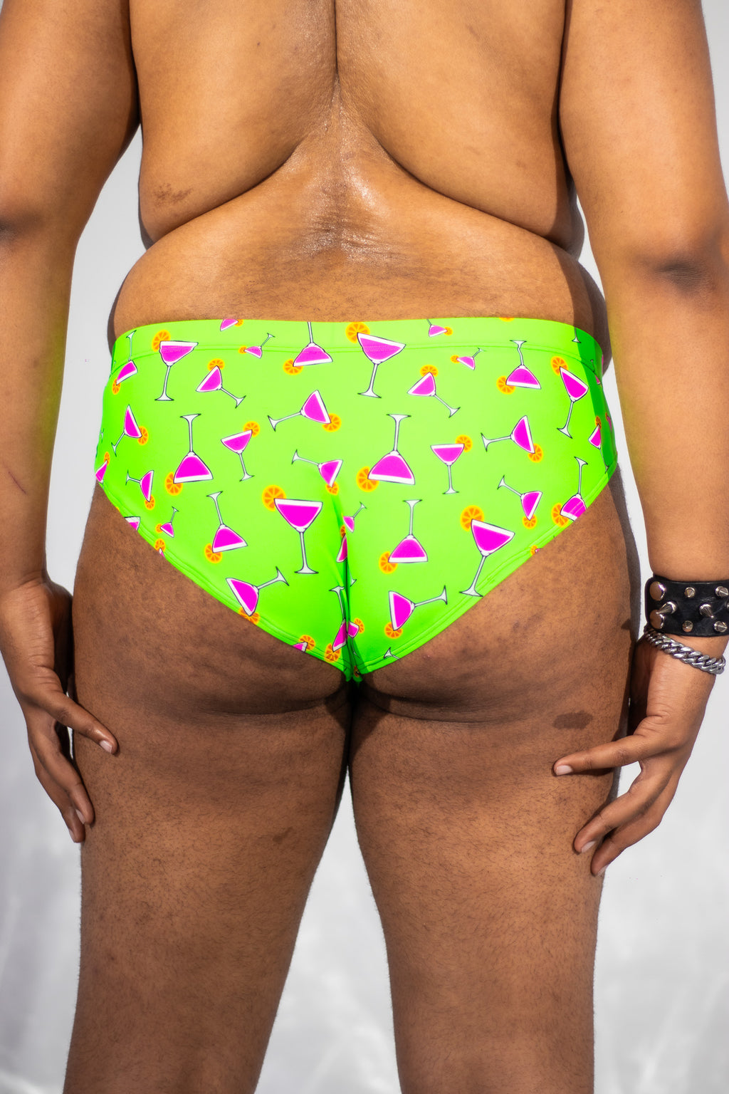 Rear view of a person wearing bright neon green bikini bottoms with a repeating pink, orange, and white cocktail glass pattern, against a light gray studio background.