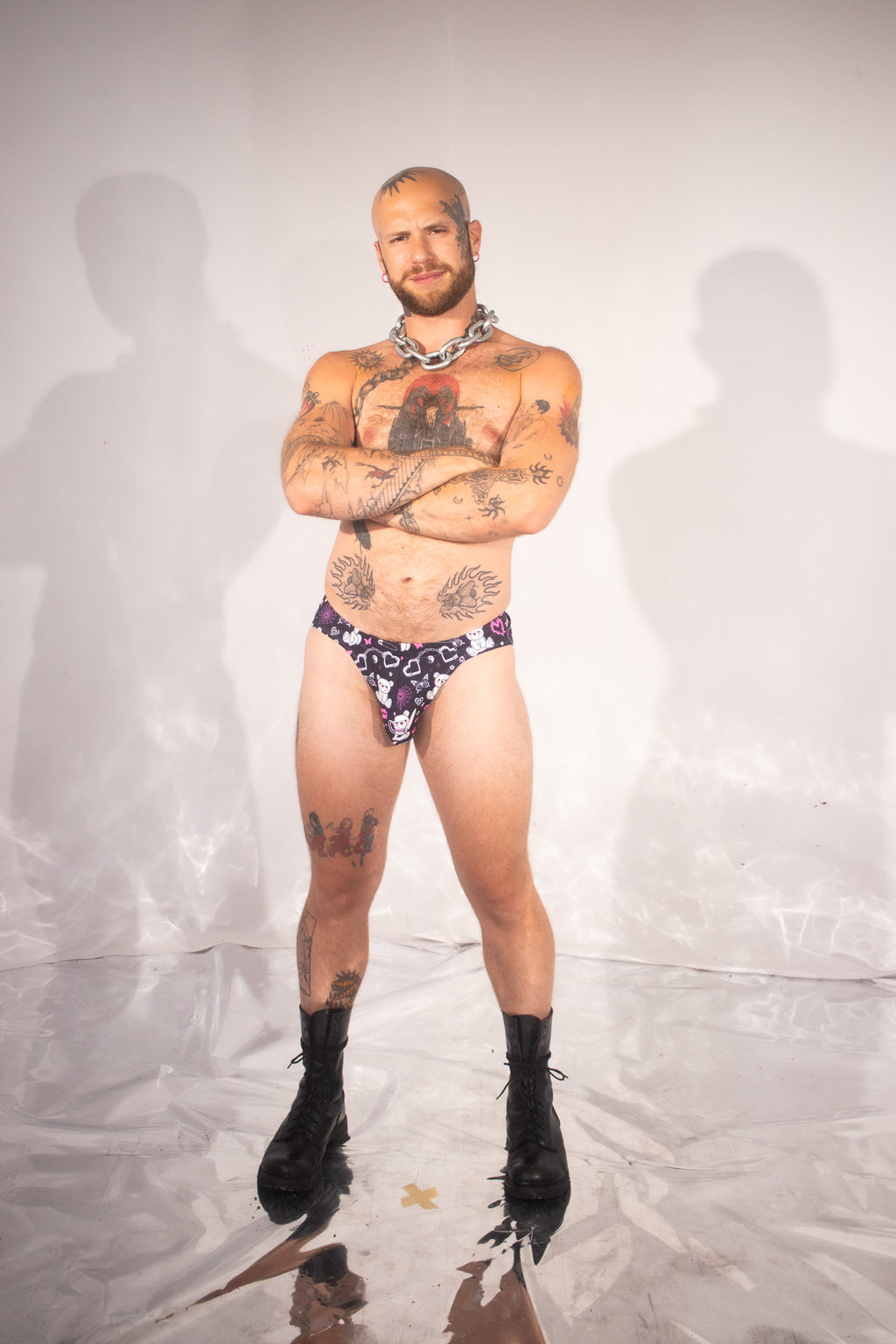 A tattooed man stands with arms crossed wearing a small patterned thong-style underwear bottom and black lace-up boots against a white studio backdrop with visible shadows.