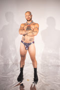 A tattooed man stands with arms crossed wearing a small patterned thong-style underwear bottom and black lace-up boots against a white studio backdrop with visible shadows.