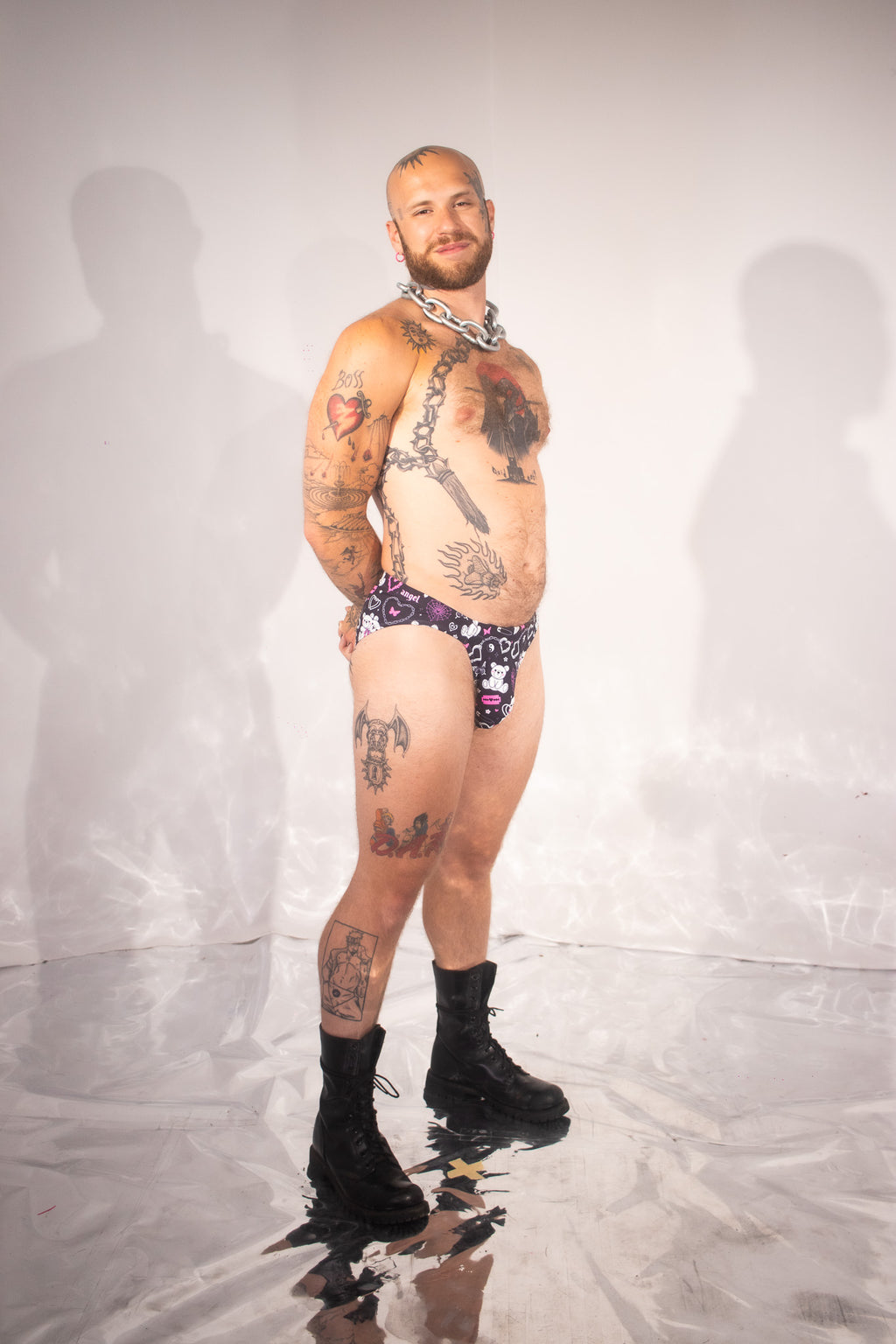 A tattooed man stands in a studio wearing patterned purple and black briefs, black boots, and a silver chain necklace against a light gray backdrop with visible shadows and a reflective floor.