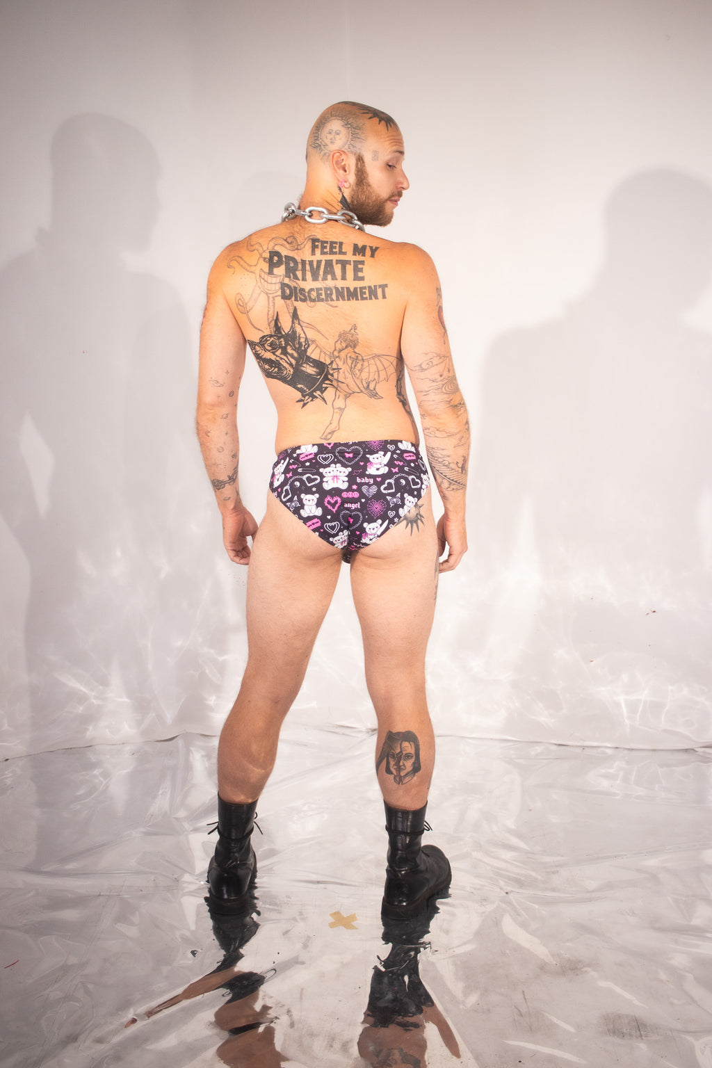 A tattooed man stands with his back to the camera wearing low-rise briefs with a purple, black, and white star pattern, paired with black boots, in front of a white studio backdrop.