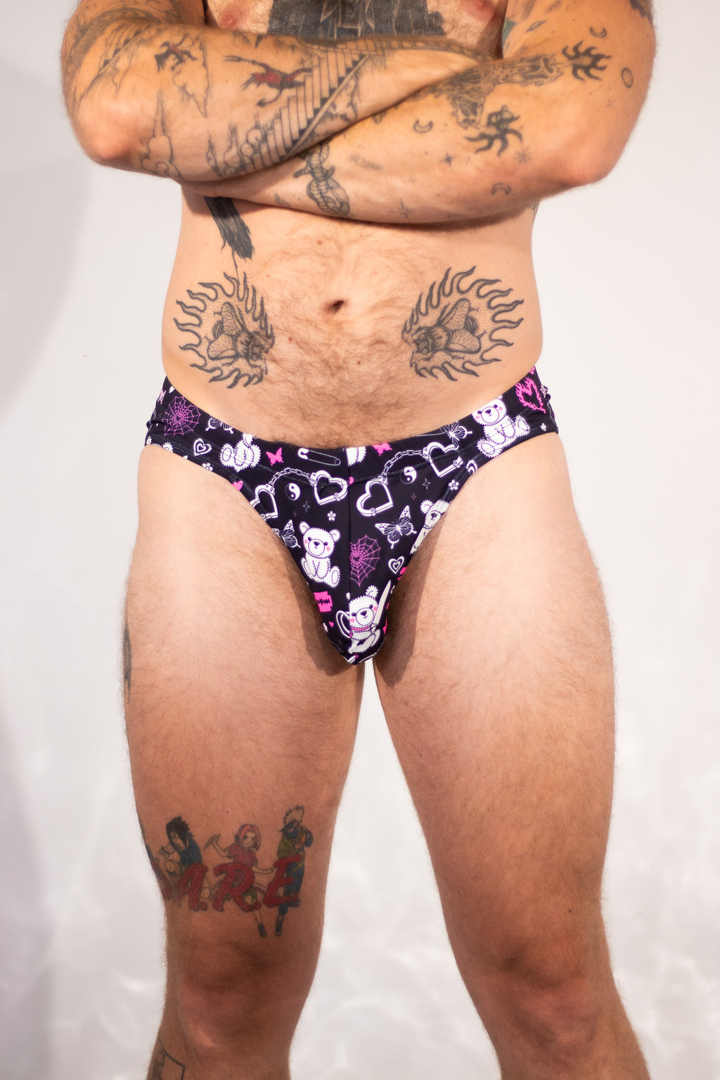 A close-up of a person wearing low-rise thong underwear with a dark purple background and a cartoon bear pattern in pink, white, and light purple. The person has visible tattoos on their arms and legs.