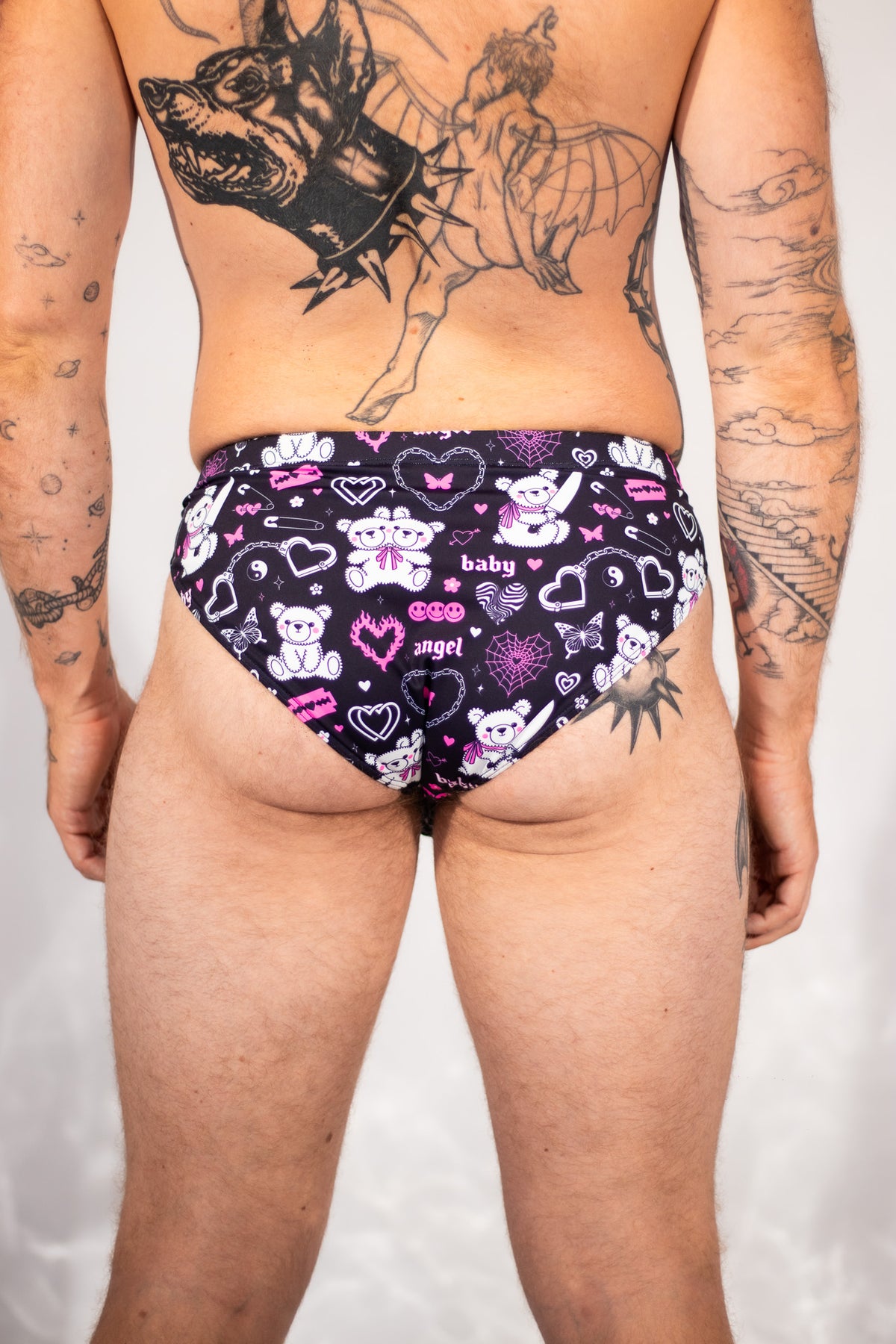Rear view of a tattooed male model wearing low-rise briefs with a black background and a pink, purple, and white teddy bear and heart pattern.