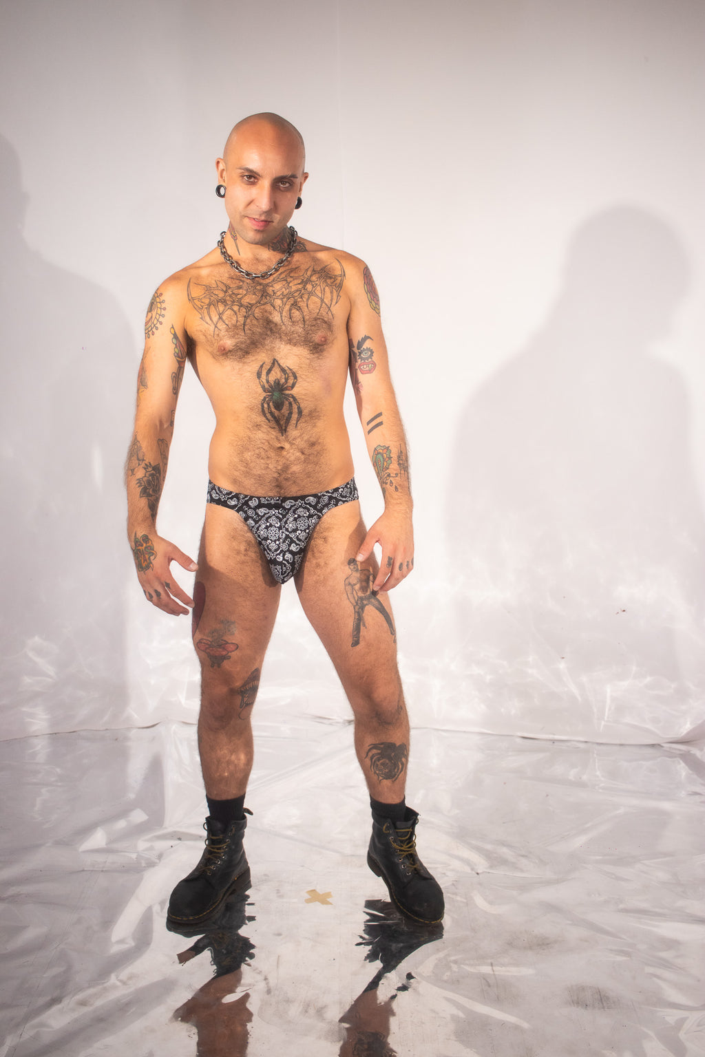 A tattooed man stands in a studio wearing a black and white patterned thong-style brief and black boots against a light gray backdrop.