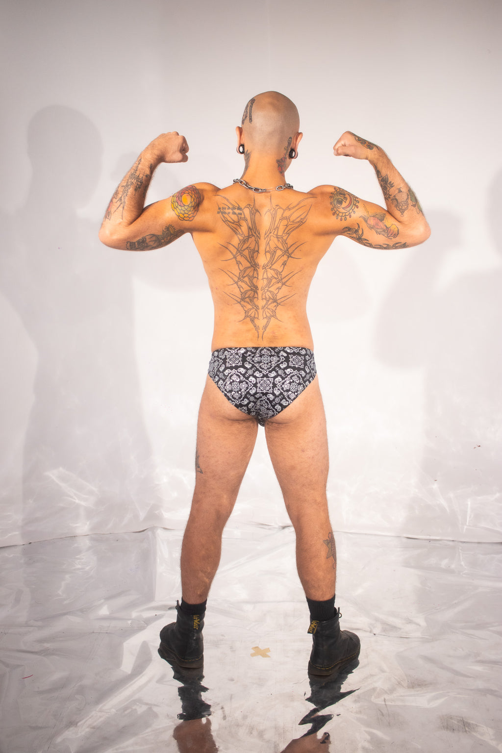 A tattooed man stands with his back to the camera, flexing his biceps while wearing low-rise patterned briefs and black boots against a white studio backdrop.