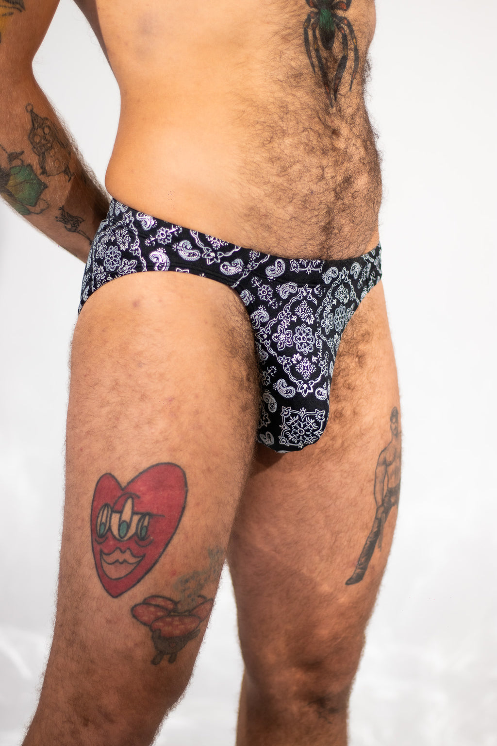 A close-up of a man's lower torso and thighs wearing low-rise briefs with a black and white paisley pattern, set against a plain white background.