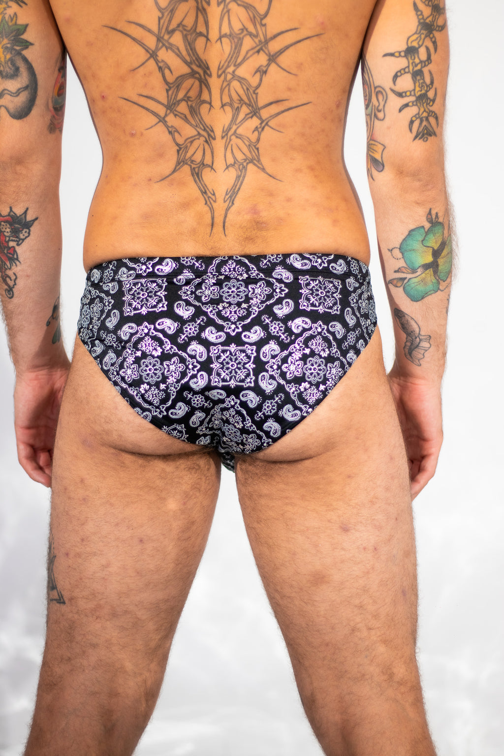 Rear view of a tattooed male model wearing low-rise briefs with a black and purple paisley pattern, set against a plain light gray background.