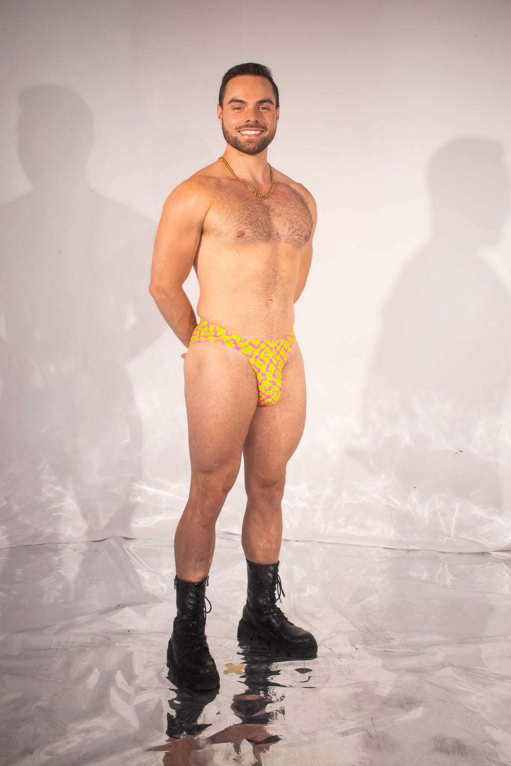 A man stands against a white studio backdrop wearing a very small yellow thong-style underwear bottom with a darker pattern, plus black combat boots. The product being highlighted is the underwear, but because a person is present the subject_type is Lifestyle.