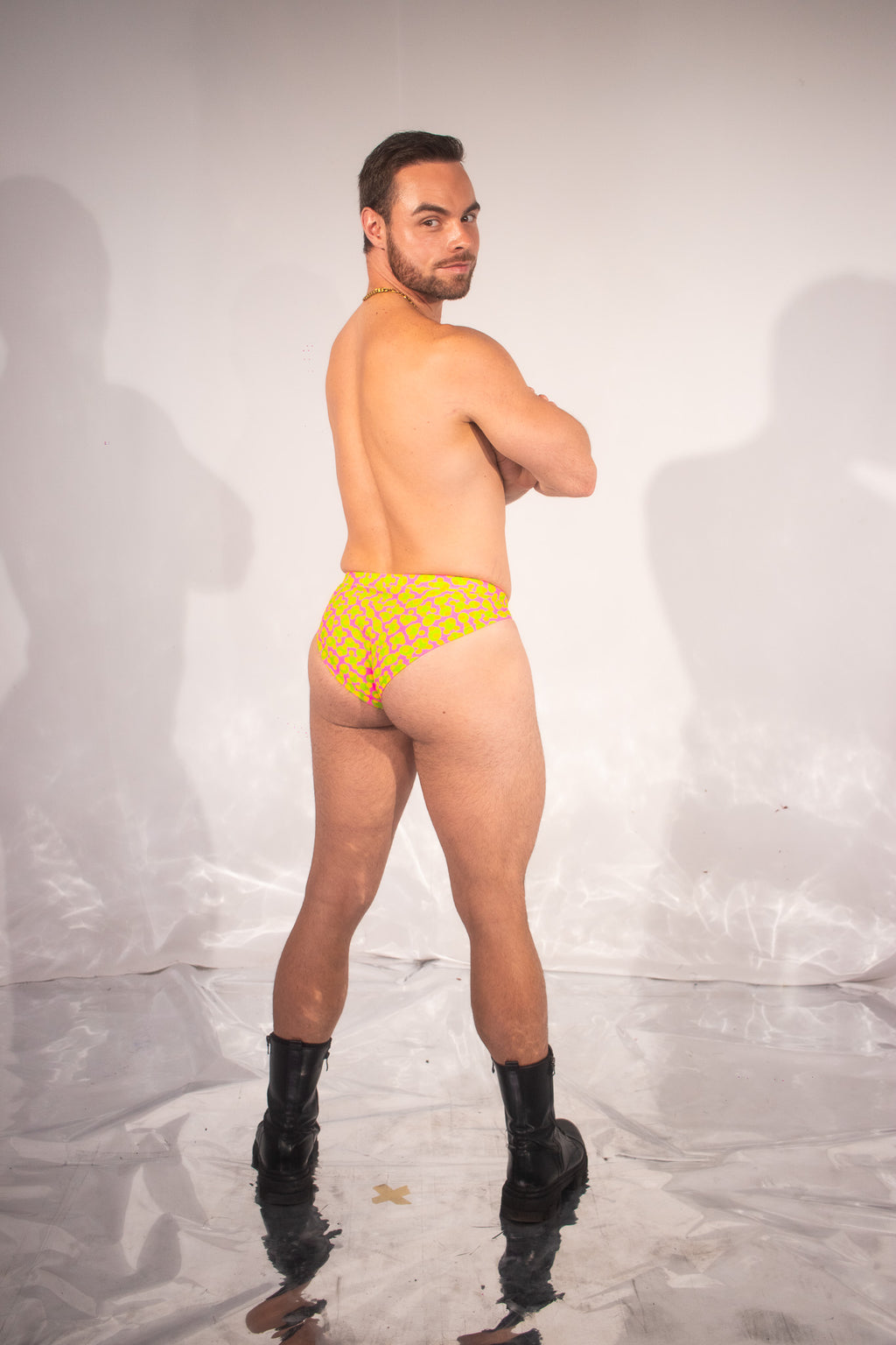 A man stands in a studio wearing bright yellow bikini-style underwear with a small repeating pattern, paired with black knee-high boots, photographed against a light gray backdrop with visible shadows.