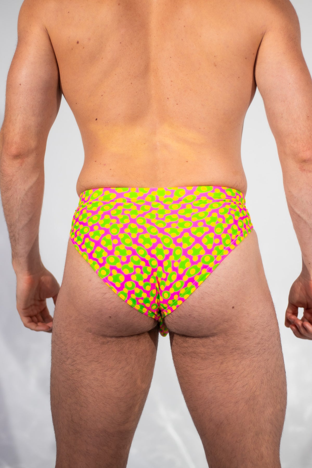 Rear view of a male model wearing low-rise briefs with a vibrant neon green and magenta geometric pattern, set against a plain light gray background.