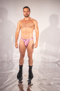 A shirtless male model wearing a pink and black patterned thong-style brief with black combat boots, standing against a white studio backdrop with visible shadows and floor reflections.