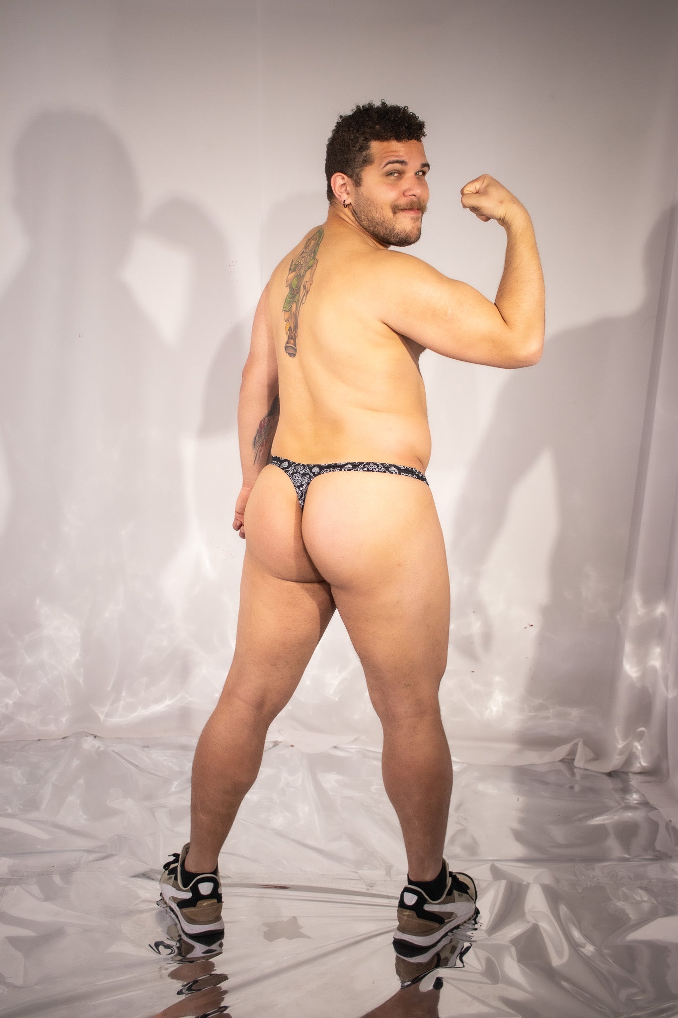 Person wearing a black and silver patterned thong and flexing against a neutral background