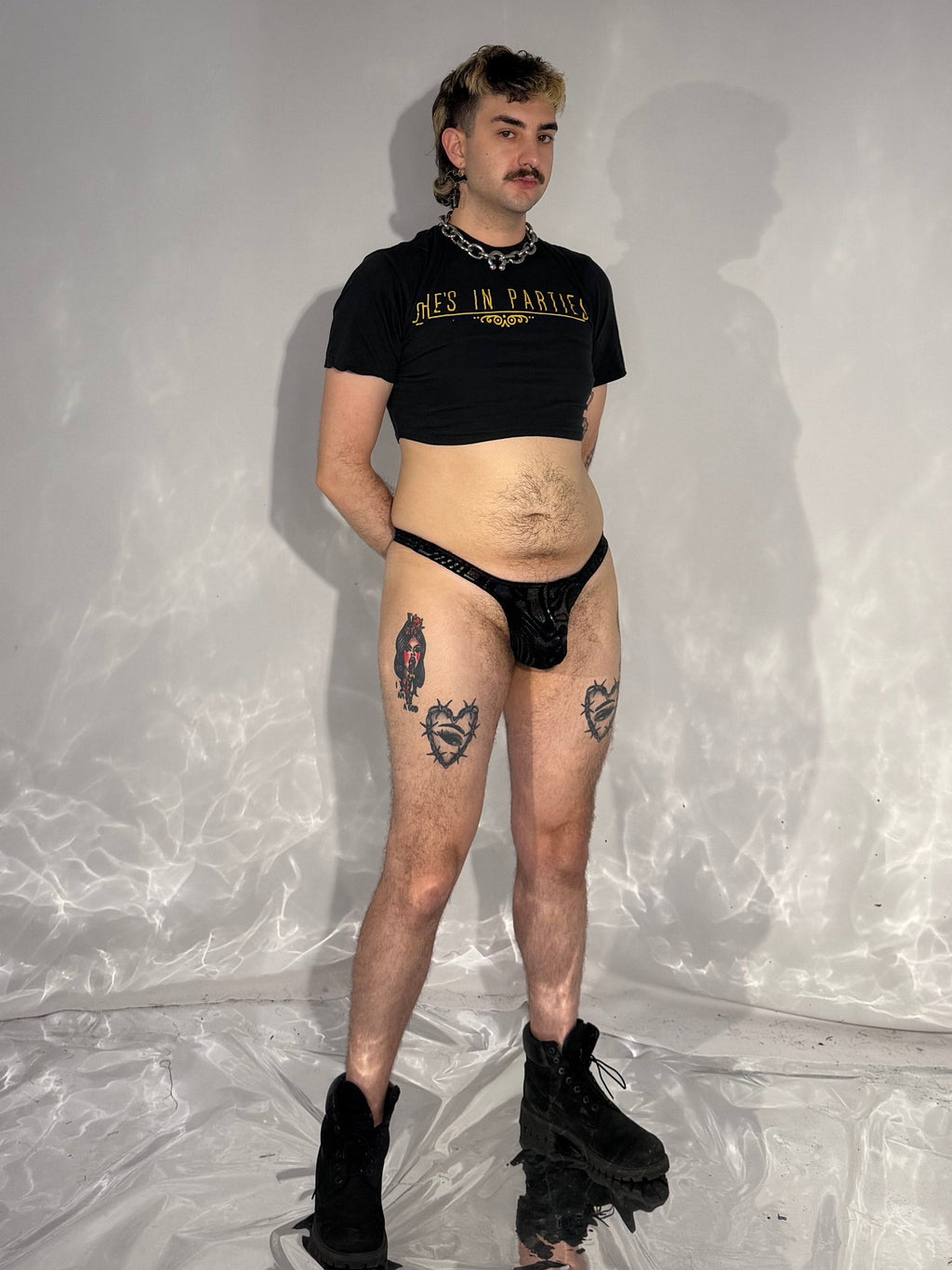 Person wearing a black t-shirt and underwear standing in a white room with reflective floor.