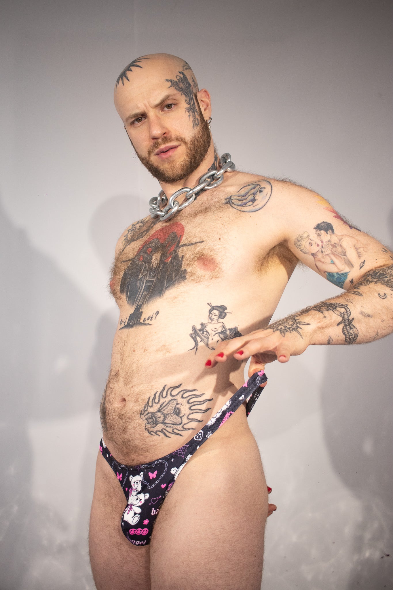 Man wearing a black, pink, and white patterned thong with tattoos on a plain background