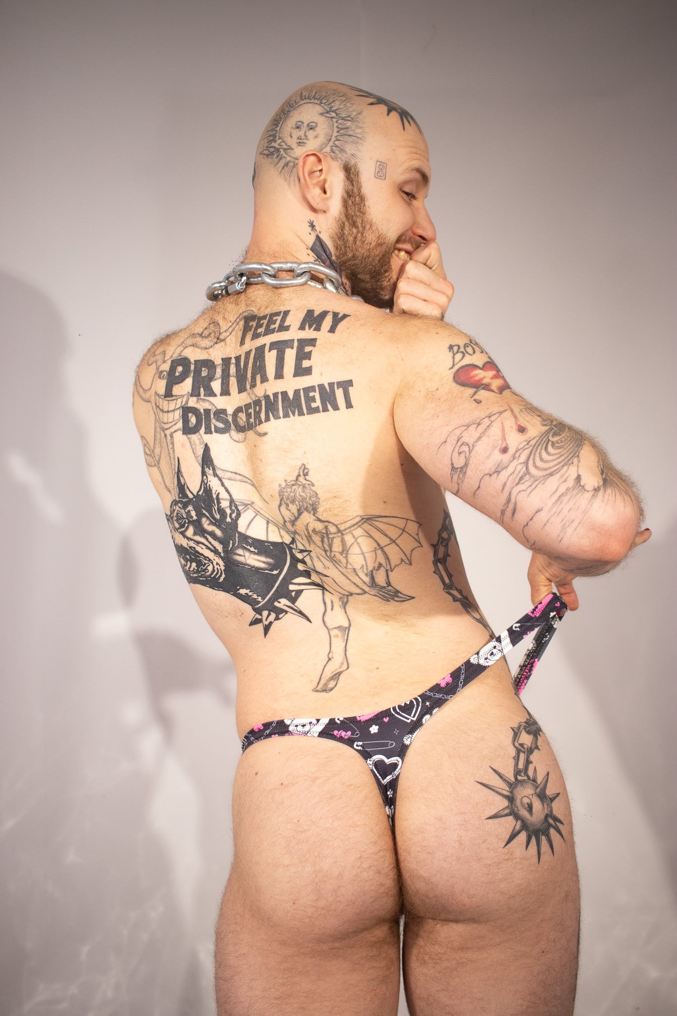 Man wearing a black, pink, and white patterned thong with tattoos on a plain background